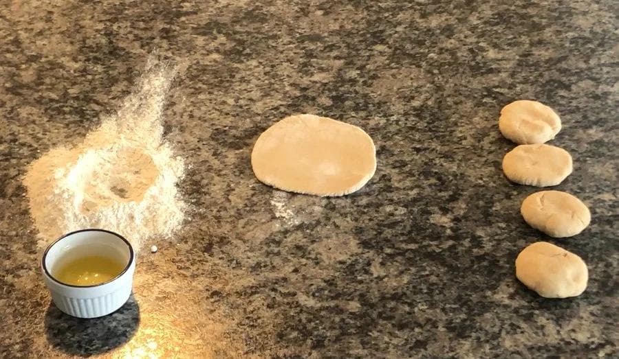 Paratha dough portions with ghee in a bowl and flour dusted on granite counter during preparation.