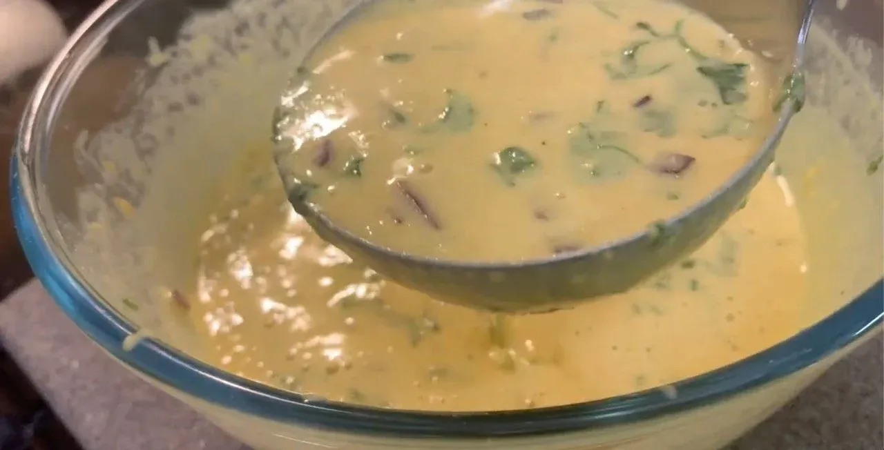 Smooth besan chilla batter in a glass bowl with green herbs sprinkled throughout, being lifted with a ladle.