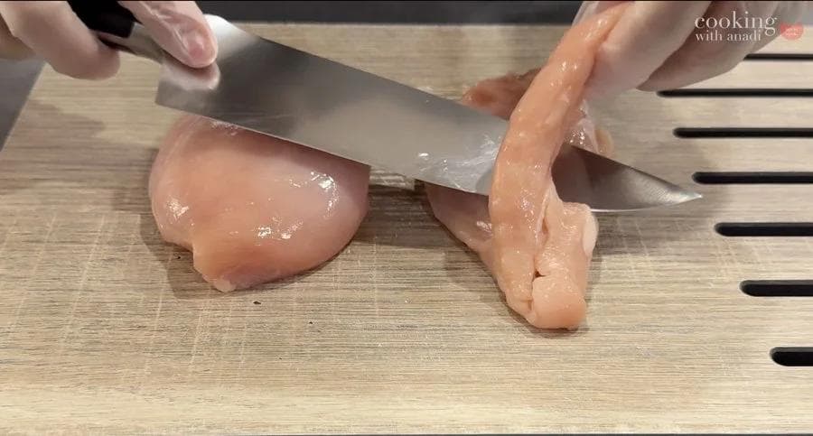 Hands pounding raw chicken breast with a meat mallet on a wooden cutting board.