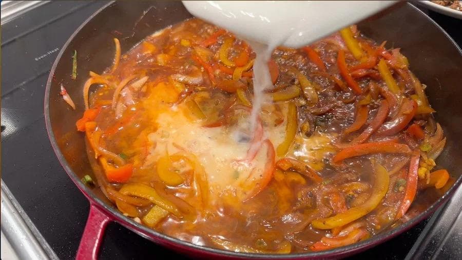 Jamaican pepper steak simmering in a red cast iron skillet with caramelized onions and peppers in rich brown sauce.