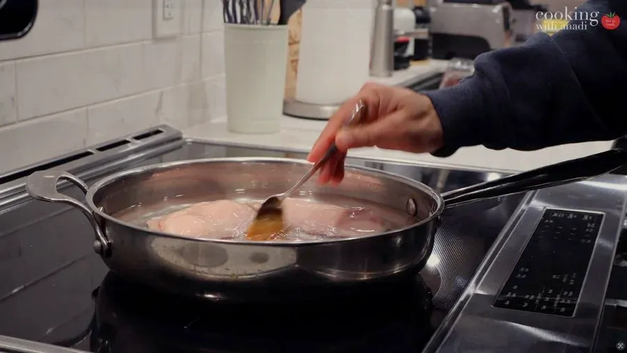Hand stirring chicken poaching in simmering water with broth in a large pot on stovetop.