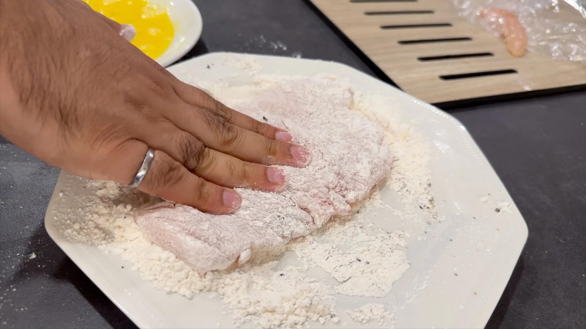 Hand dredging raw chicken cutlet in seasoned flour on a white plate during schnitzel preparation.