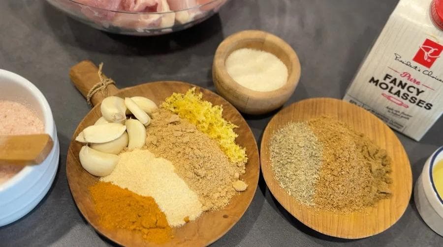 Spice blend ingredients for satay marinade arranged on wooden plate: turmeric, cinnamon, cumin, sliced garlic, and molasses.
