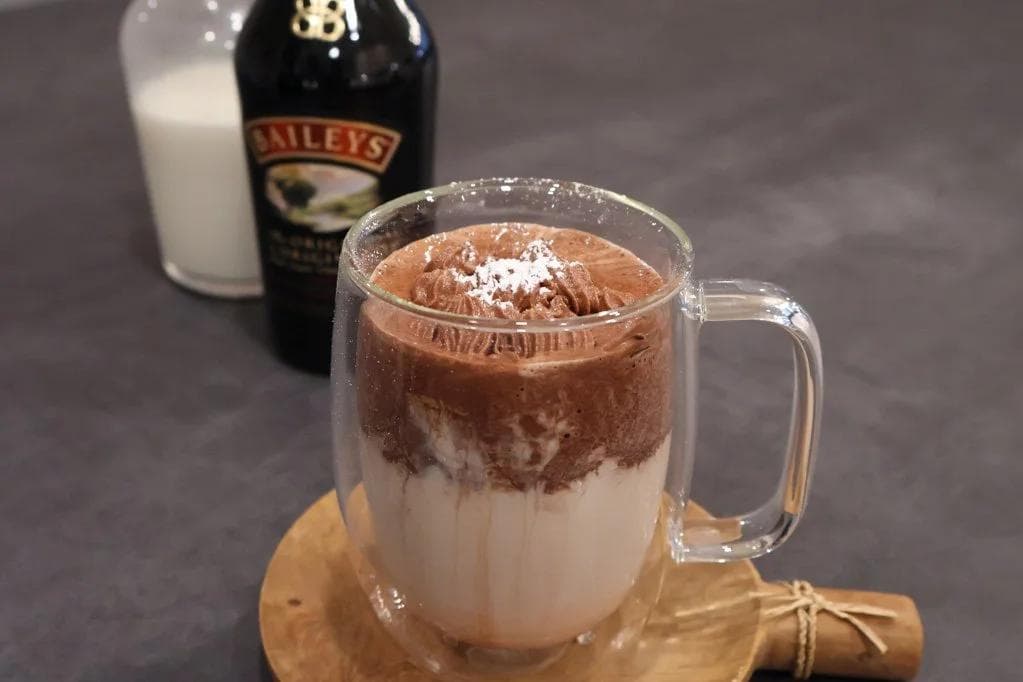 Whipped Baileys hot chocolate in a glass mug with cocoa powder on top, milk below, next to a Baileys bottle and glass of milk.