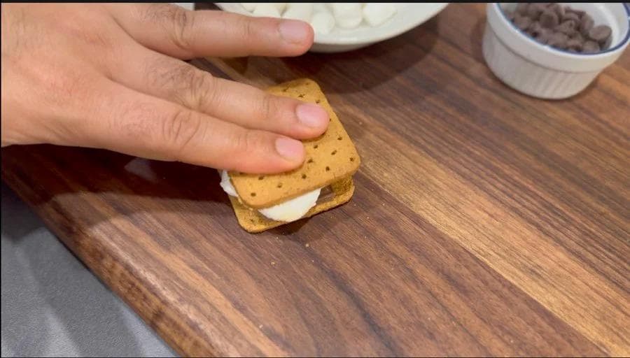 Hand pressing a graham cracker onto melted chocolate and marshmallow filling on wooden surface.