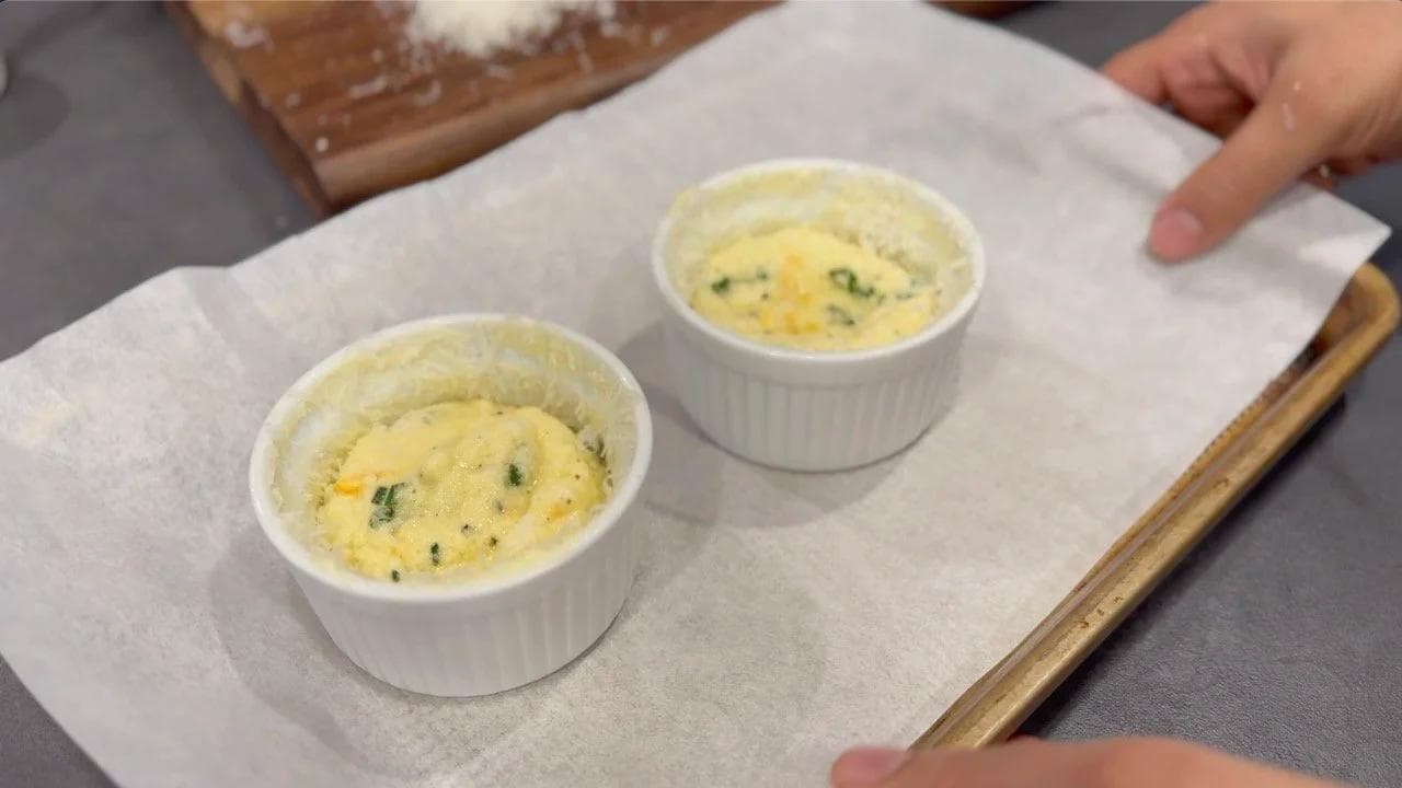 Two baked cheesy egg soufflés in white ramekins on parchment paper, risen and golden on top.