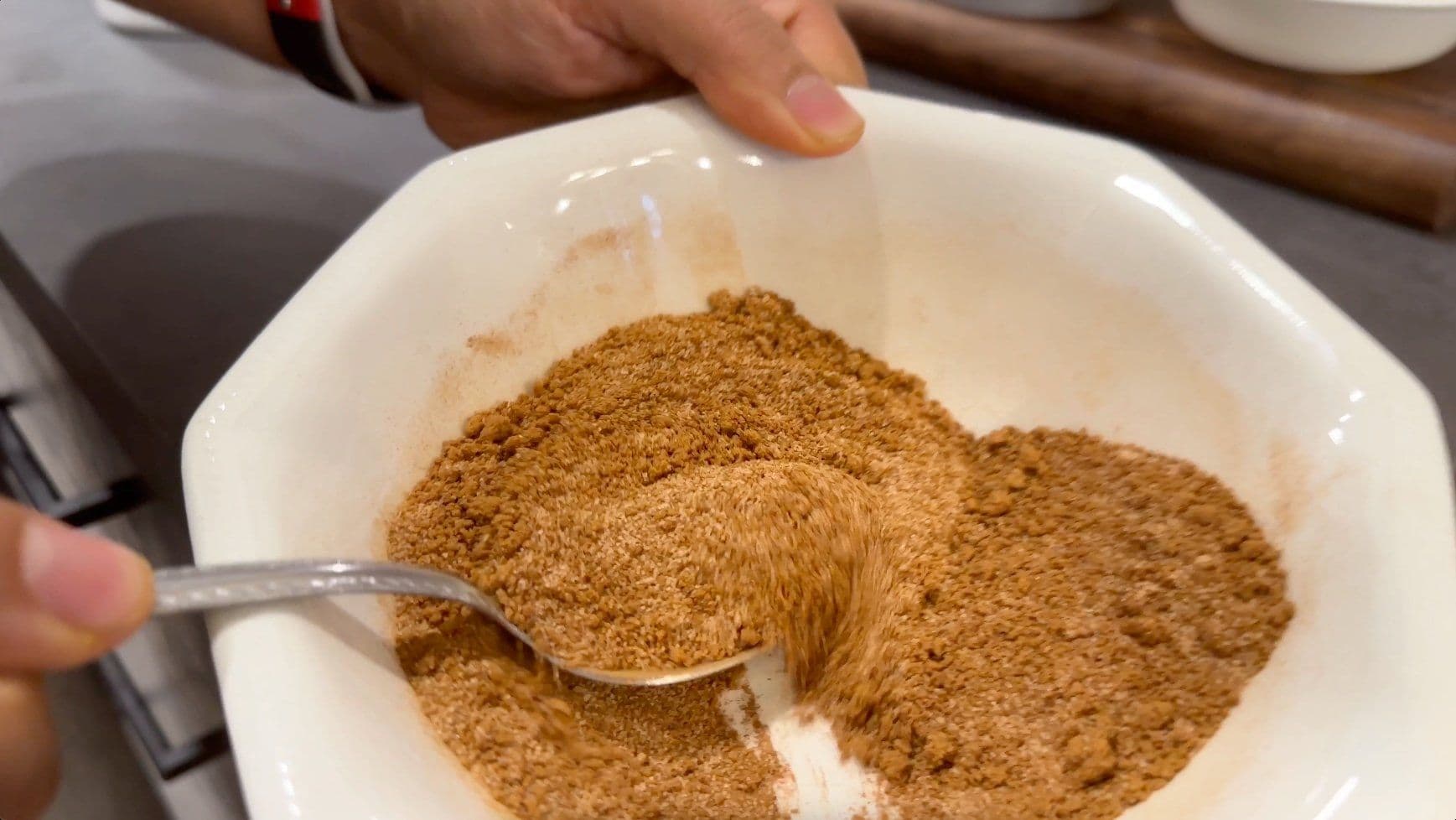 Cinnamon sugar mixture being scooped with a spoon in a white bowl for French toast casserole topping.
