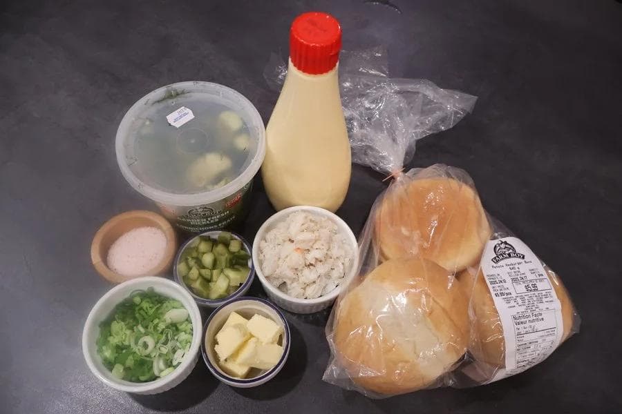 Ingredients for crab salad sandwiches laid out on a dark surface: canned crab, bread rolls, mayonnaise, pickles, green onions, butter, and seasonings.