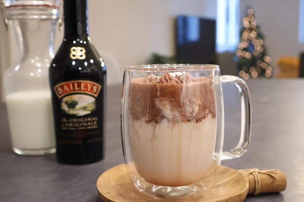 Baileys hot chocolate with whipped cream topping in a glass mug, next to Baileys bottle and milk glass.