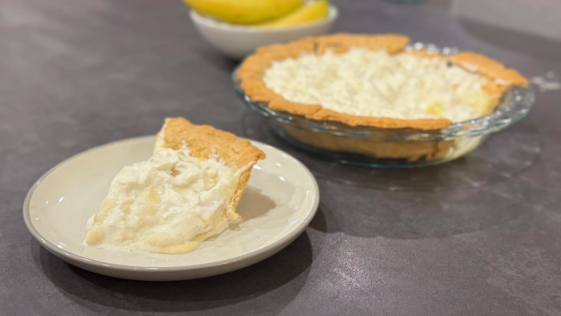 Slice of banana cream pie with golden crust and thick whipped cream filling on a white plate, whole pie in background.