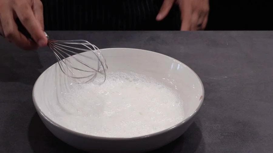 Whisk being used to mix wet batter in a white bowl for breading chicken nuggets.