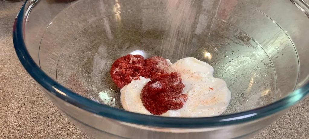 Chicken marinating in yogurt, tomato paste, and spices in a glass bowl.