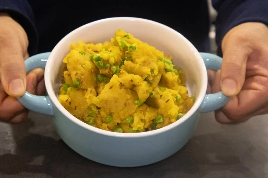 Hands holding a blue bowl of golden yellow mashed potatoes with green peas stirred throughout.