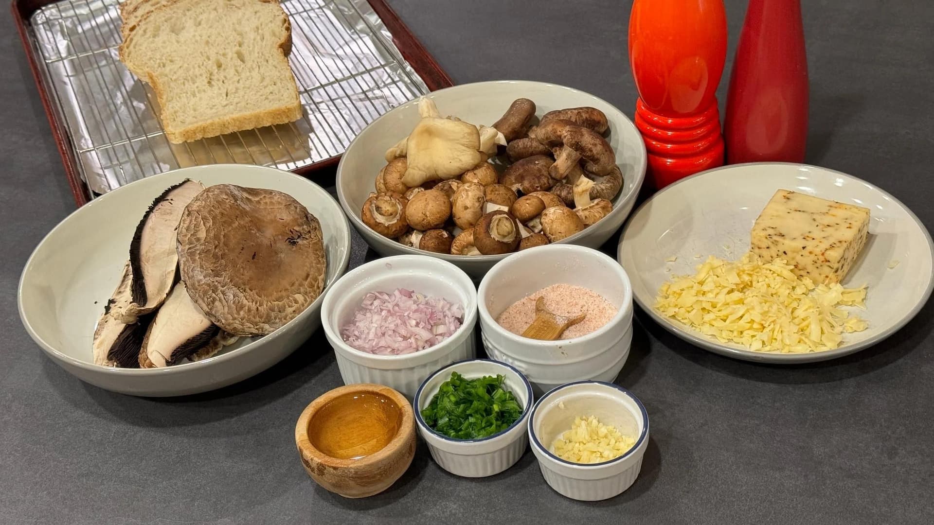 Ingredients laid out for mushrooms on toast: mixed mushrooms, bread, cheese, shallots, garlic, breadcrumbs, and seasonings in white bowls.