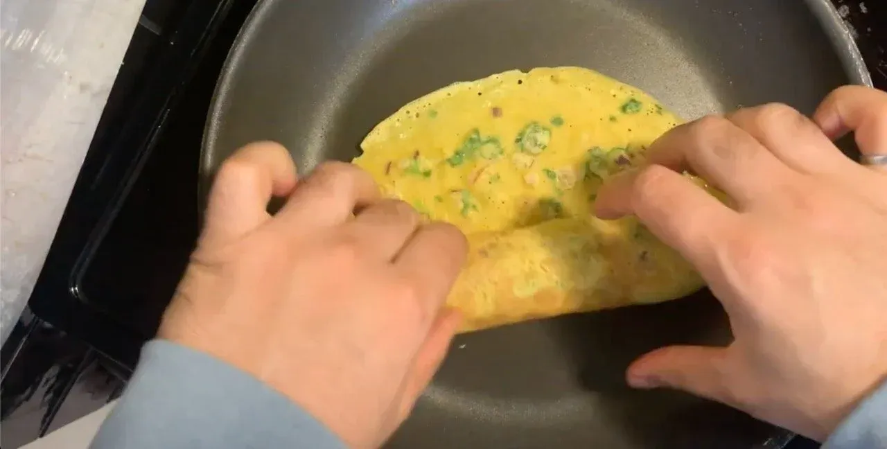 Hands flipping a yellow besan chilla pancake with green herbs in a dark skillet.