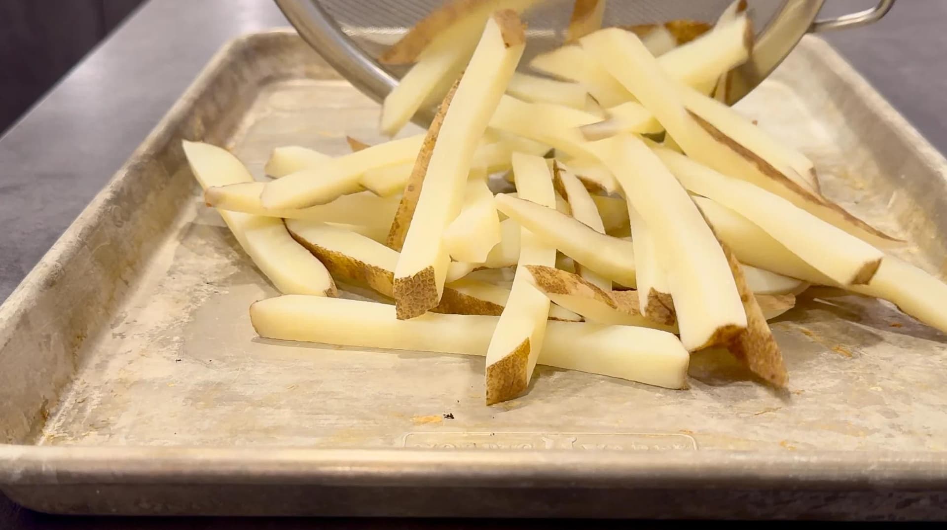 Parboiled potato fries draining on parchment paper in a metal tray before air frying.