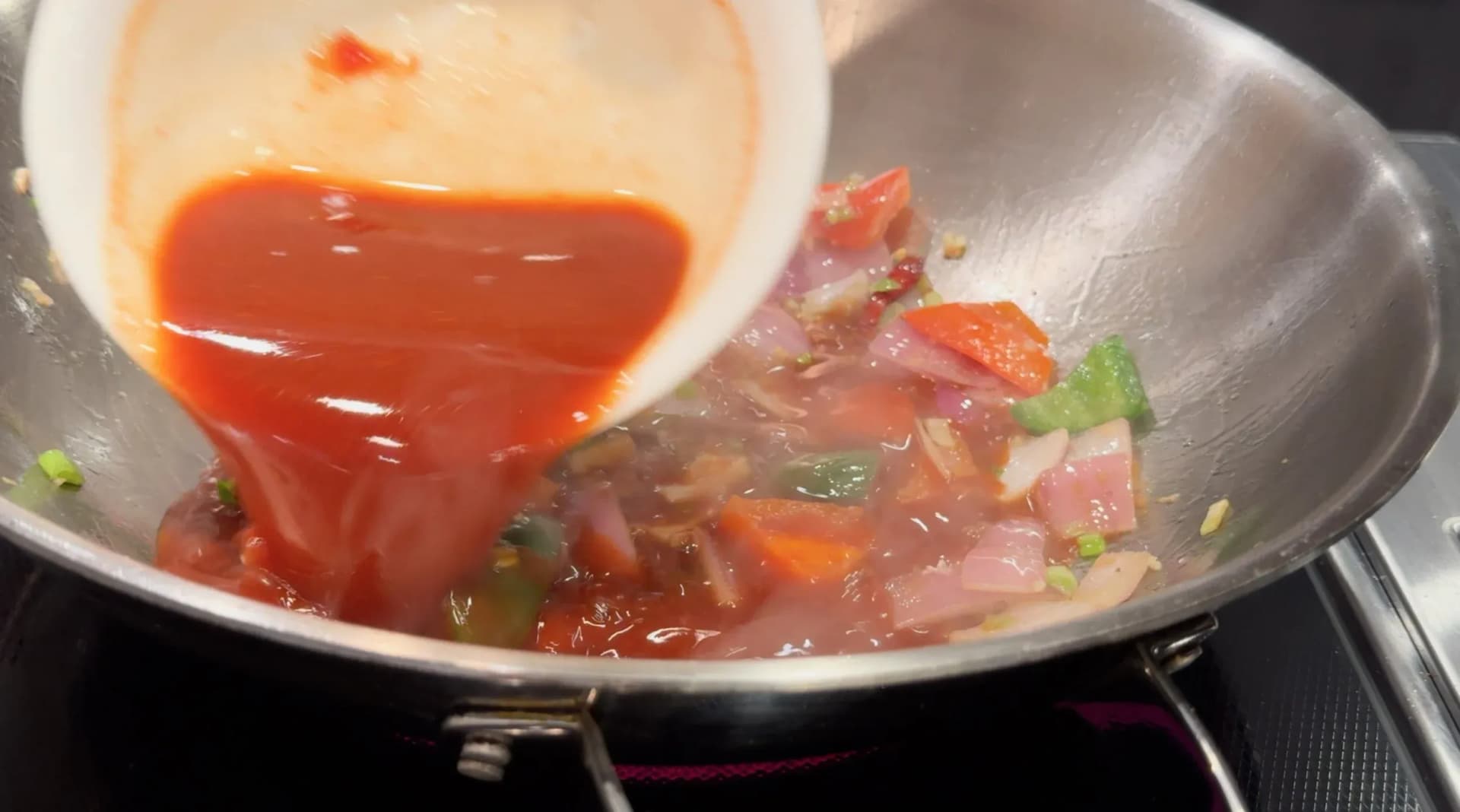 Red tomato-based sauce being poured from a white bowl into a wok with sautéed onions and green peppers.