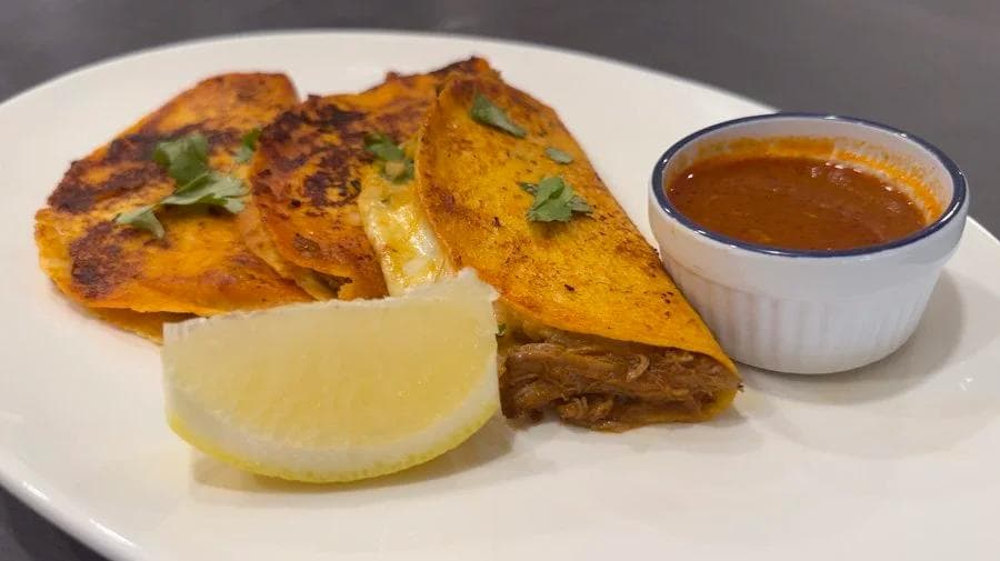 Crispy birria tacos on a white plate with a lime wedge and small bowl of consomé sauce for dipping.