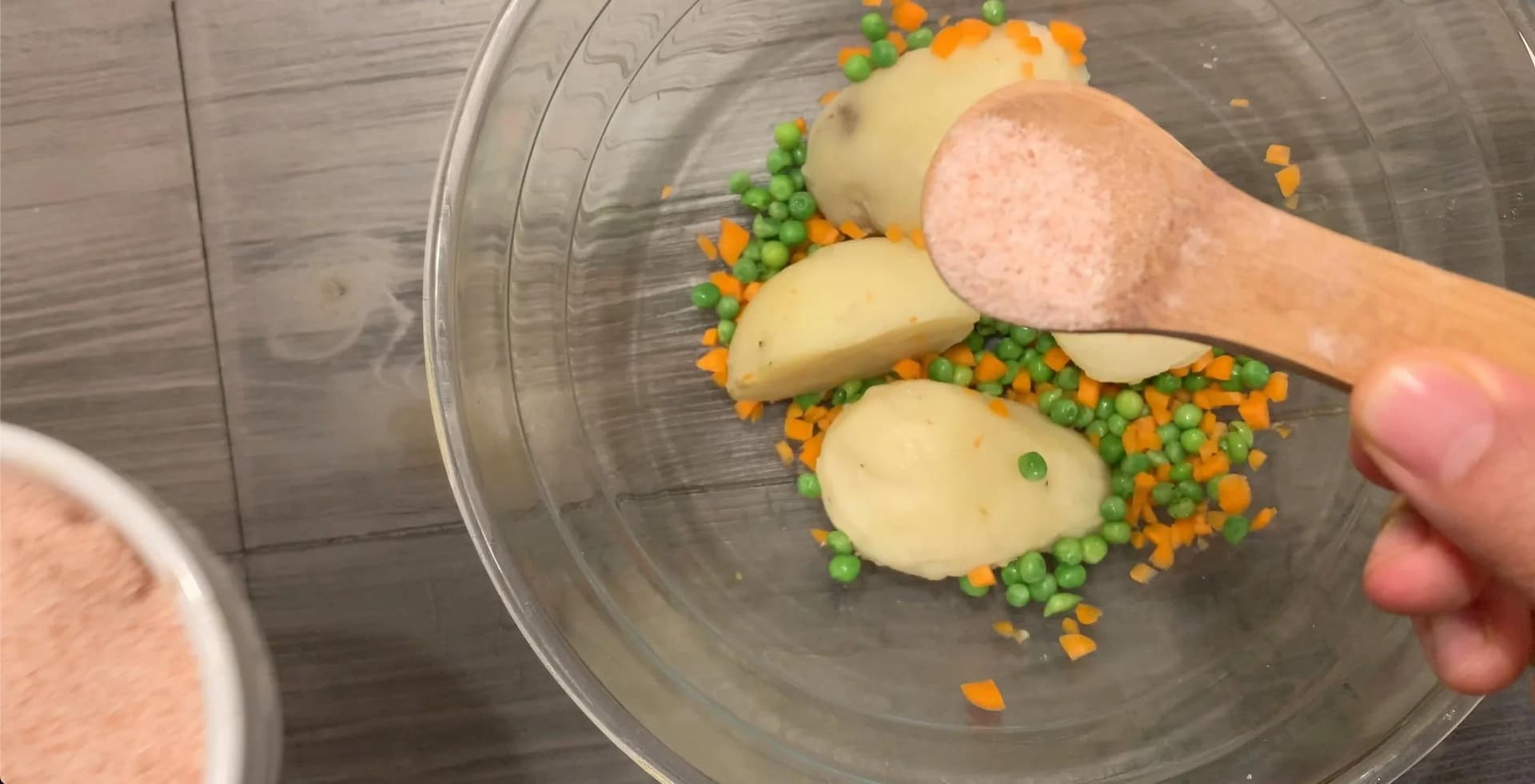 Mashed potato mixture with peas and carrots being mixed with a wooden spoon in a glass bowl