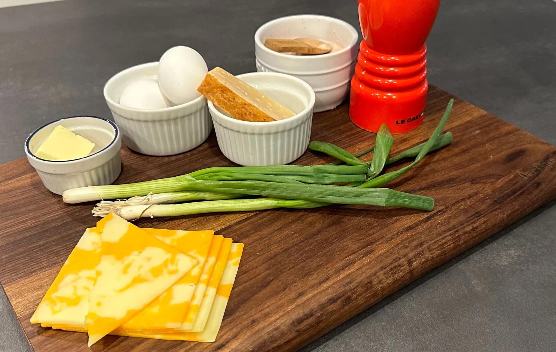 Ingredients for cheesy egg soufflé arranged on wooden board: eggs, cheddar cheese slices, butter, green onions, and seasonings.