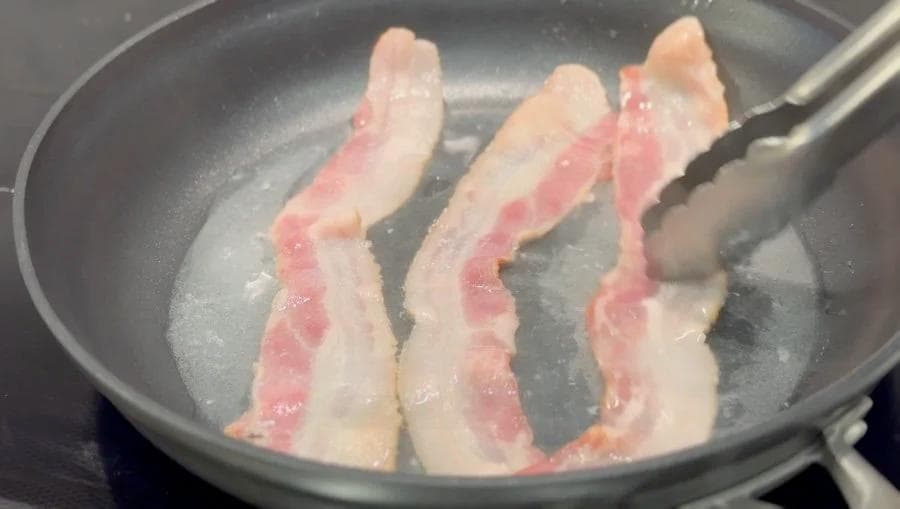 Three strips of bacon cooking in a dark skillet with a spatula lifting one strip.