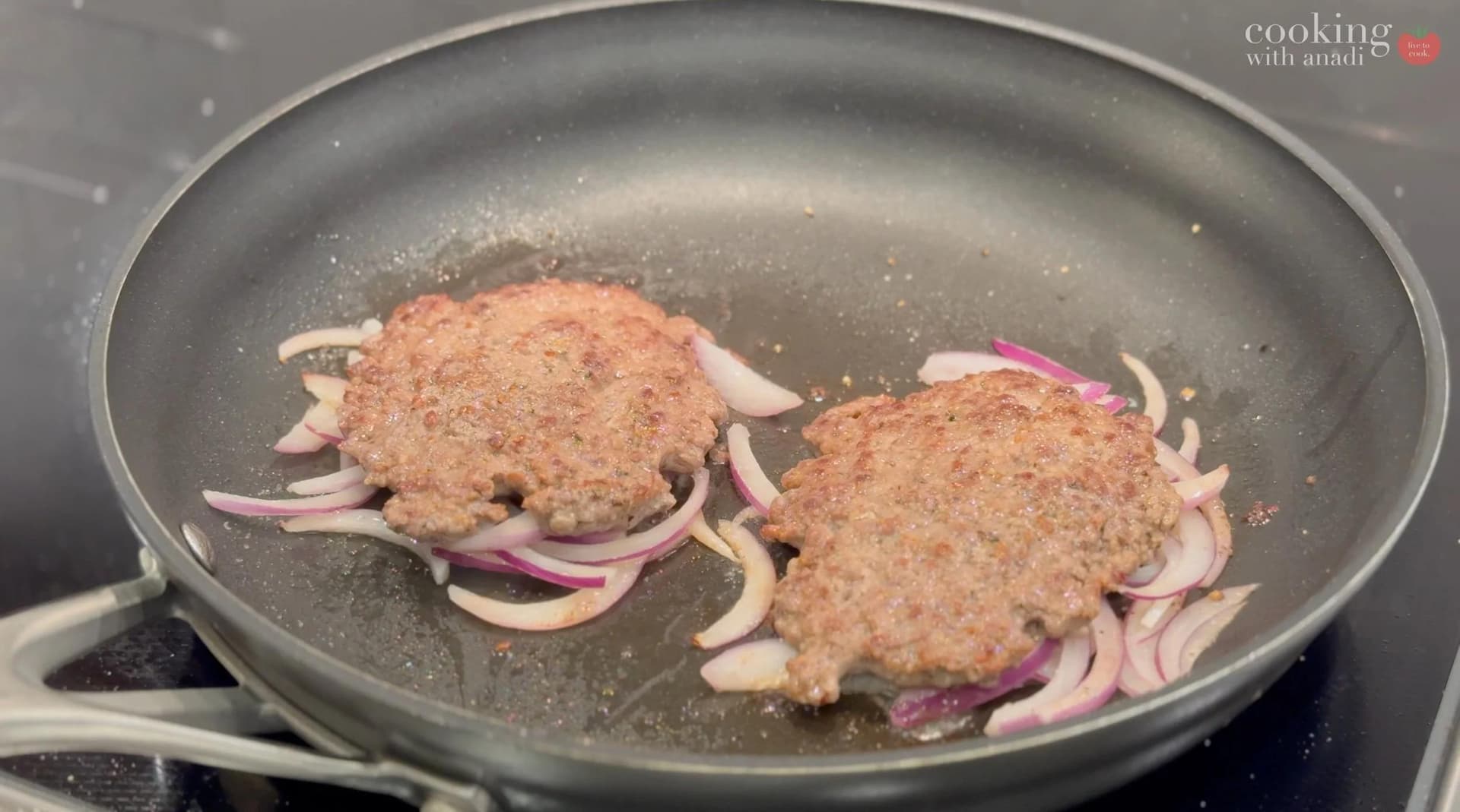 Two smash burger patties with sliced red onions cooking in a nonstick skillet.