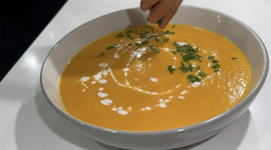 Creamy carrot ginger soup garnished with coconut milk swirl, chopped chives, and fresh herbs in a white bowl.