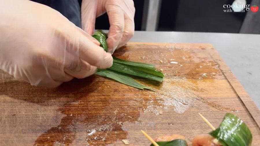 Gloved hands wrapping marinated chicken in pandan leaves on a wooden cutting board, with toothpick-secured parcels nearby.