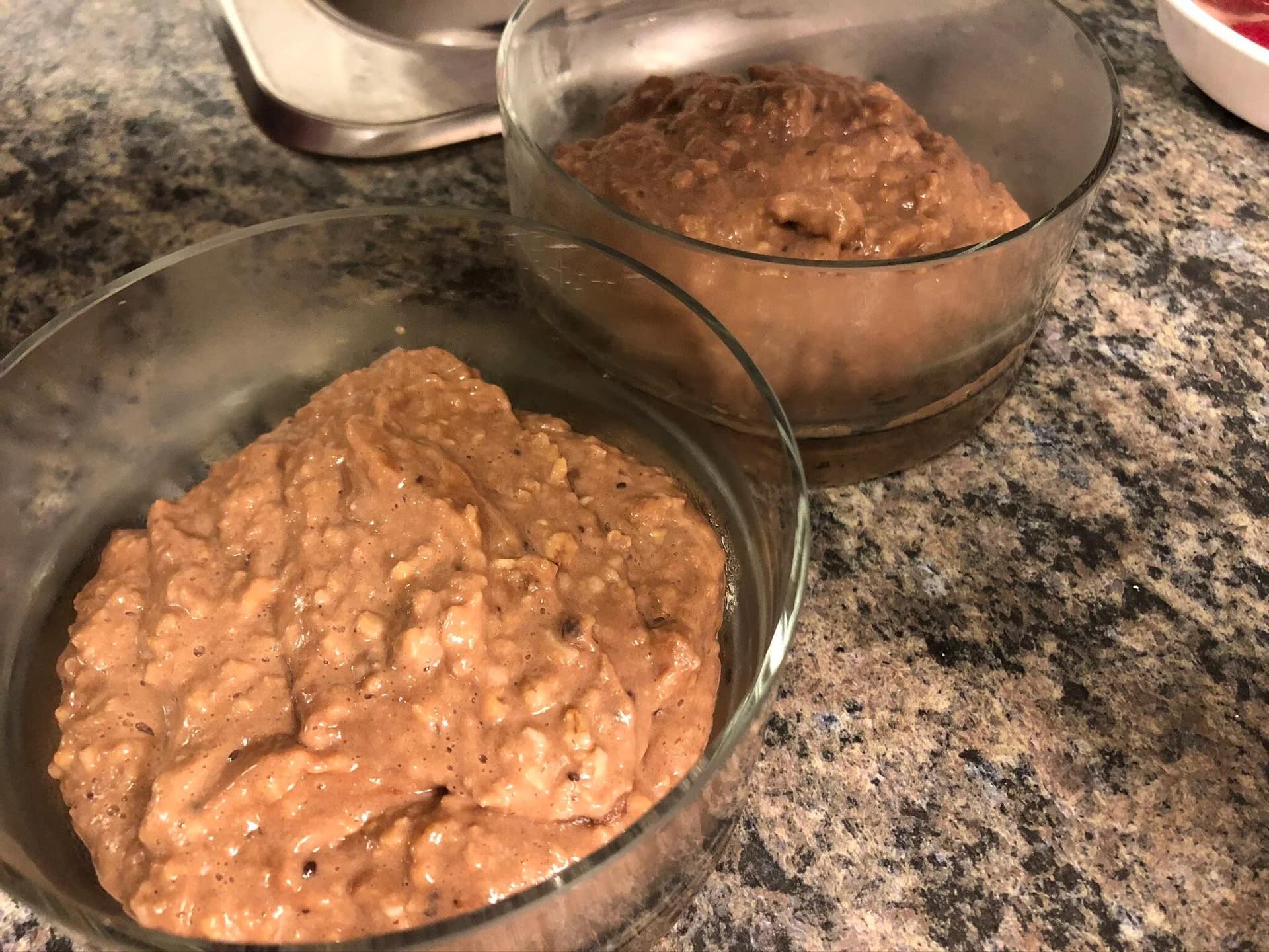 Chocolate protein oatmeal mixture in two glass bowls on a granite countertop, ready to be served.