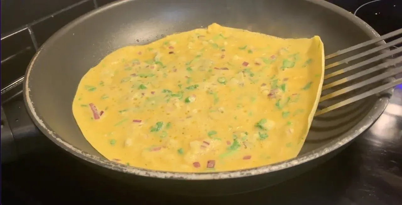 Golden besan chilla cooking in a pan with green onions and red peppers visible on top.
