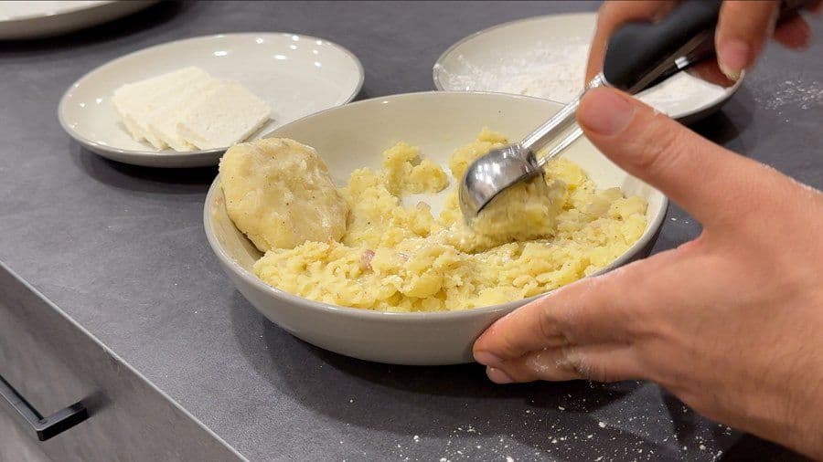 Hands shaping mashed potatoes with a cheese-filled center into a patty over a bowl of potato mixture.