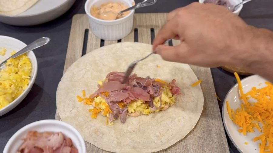 Hands folding a breakfast burrito with scrambled eggs, ham, and cheese on a flour tortilla.