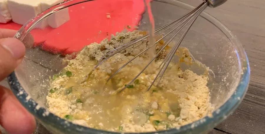 Whisking besan flour batter with spices and herbs in a glass bowl for paneer pakora.