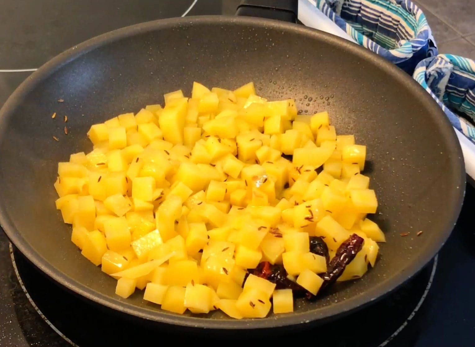 Diced potatoes cooking in a dark nonstick skillet with cumin seeds and dried red chilies, coated in turmeric.