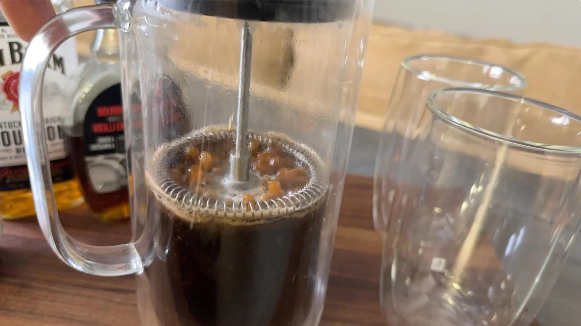 French press with brewed dark coffee steeping, surrounded by empty glasses and liquor bottles on wooden surface.