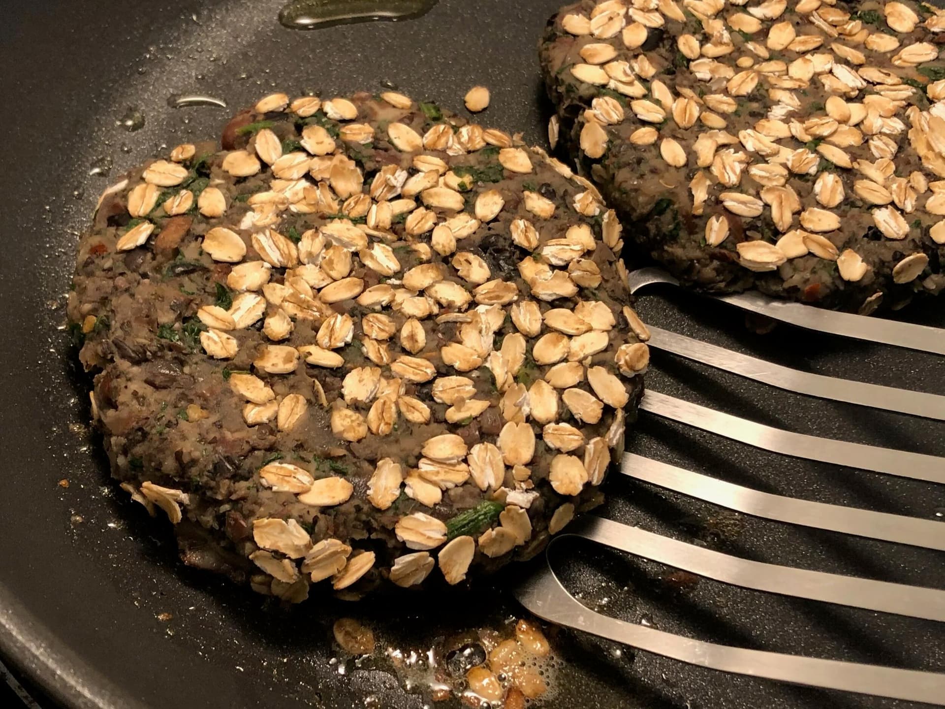 Two vegetarian bean patties topped with sliced almonds cooking in a skillet with a spatula.