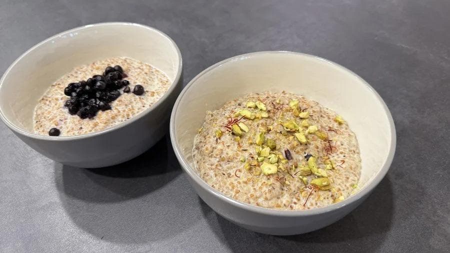 Two bowls of creamy cracked wheat porridge topped with black currants and pistachios on a gray surface.