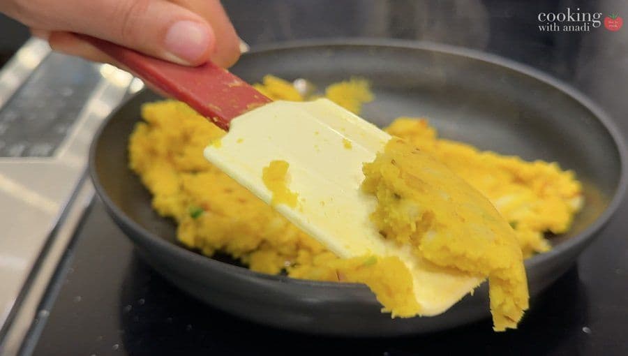 Butter being added to cooled yellow mashed potatoes in a dark pan with a spatula.