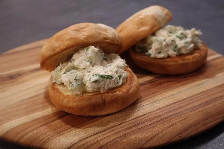 Two crab salad sandwiches on toasted buns with fresh herbs, served on a wooden cutting board.