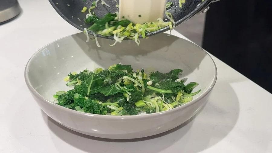 Sautéed leeks and kale with green onions being transferred from a skillet to a white bowl.