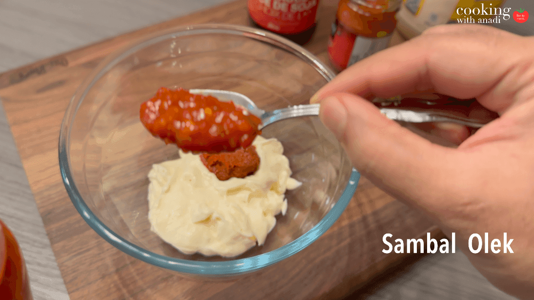 Spoon of red sambal olek being added to creamy mayonnaise in a glass bowl for curry mayo.