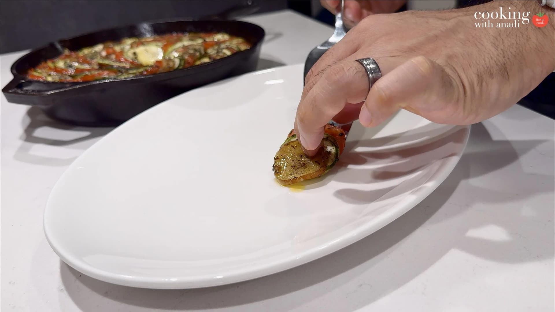 Hand placing a slice of baked ratatouille onto a white oval platter, with a cast iron skillet of piperade in the background.