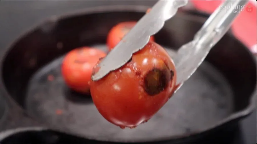 Charred tomato held with tongs over a hot cast iron pan with additional blackened tomatoes visible in the background.