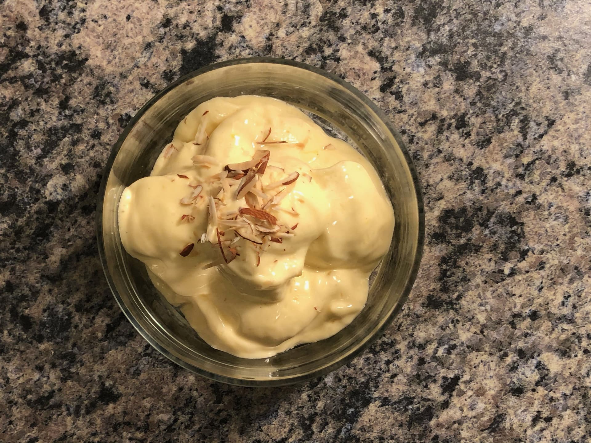Vanilla protein ice cream topped with sliced almonds in a glass bowl on a granite countertop.