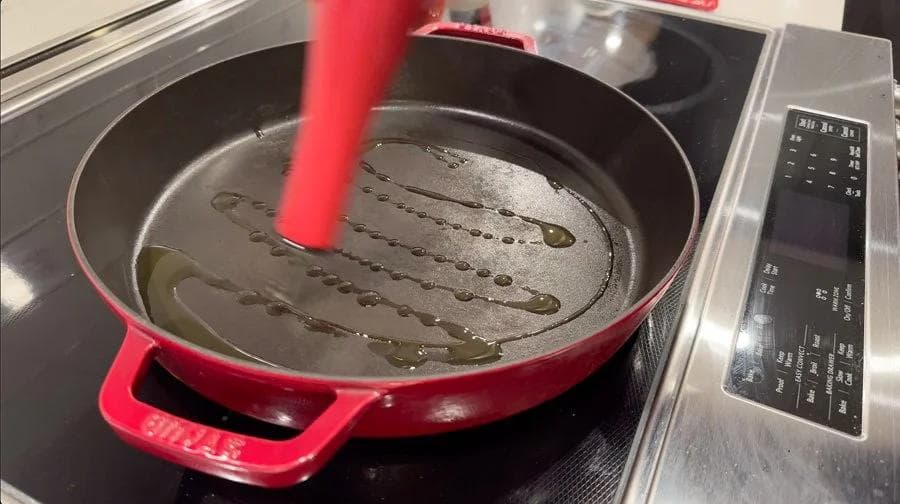 Red cast-iron pan with hot olive oil and sliced peppers and onions being added over medium-high heat on stovetop.