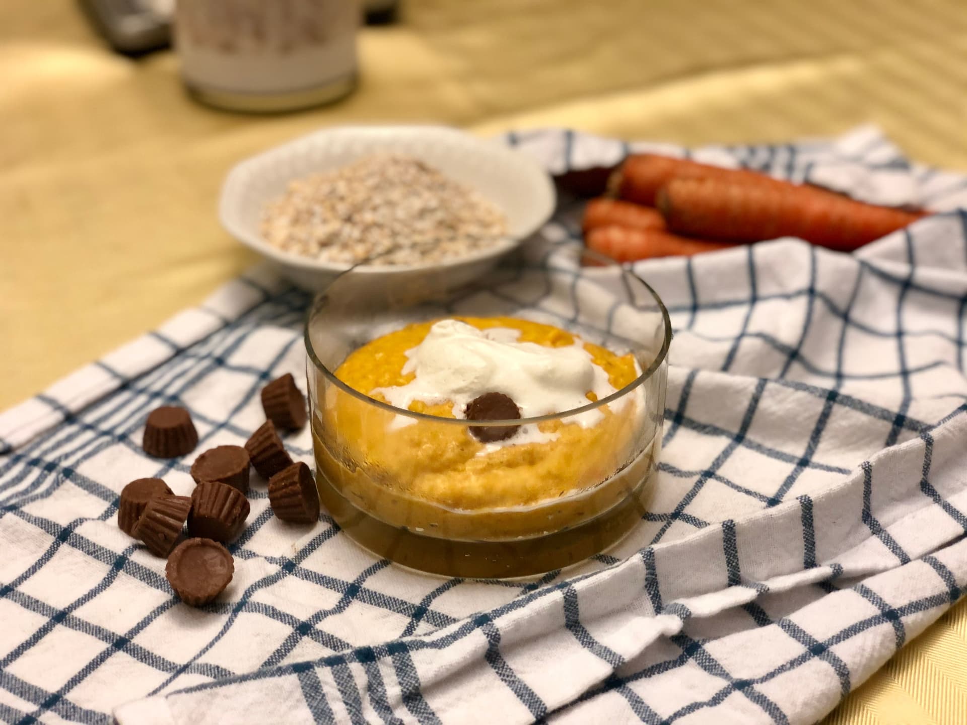 Reese's carrot cheesecake protein oatmeal in a glass bowl topped with whipped cream and chocolate chips, surrounded by ingredients.