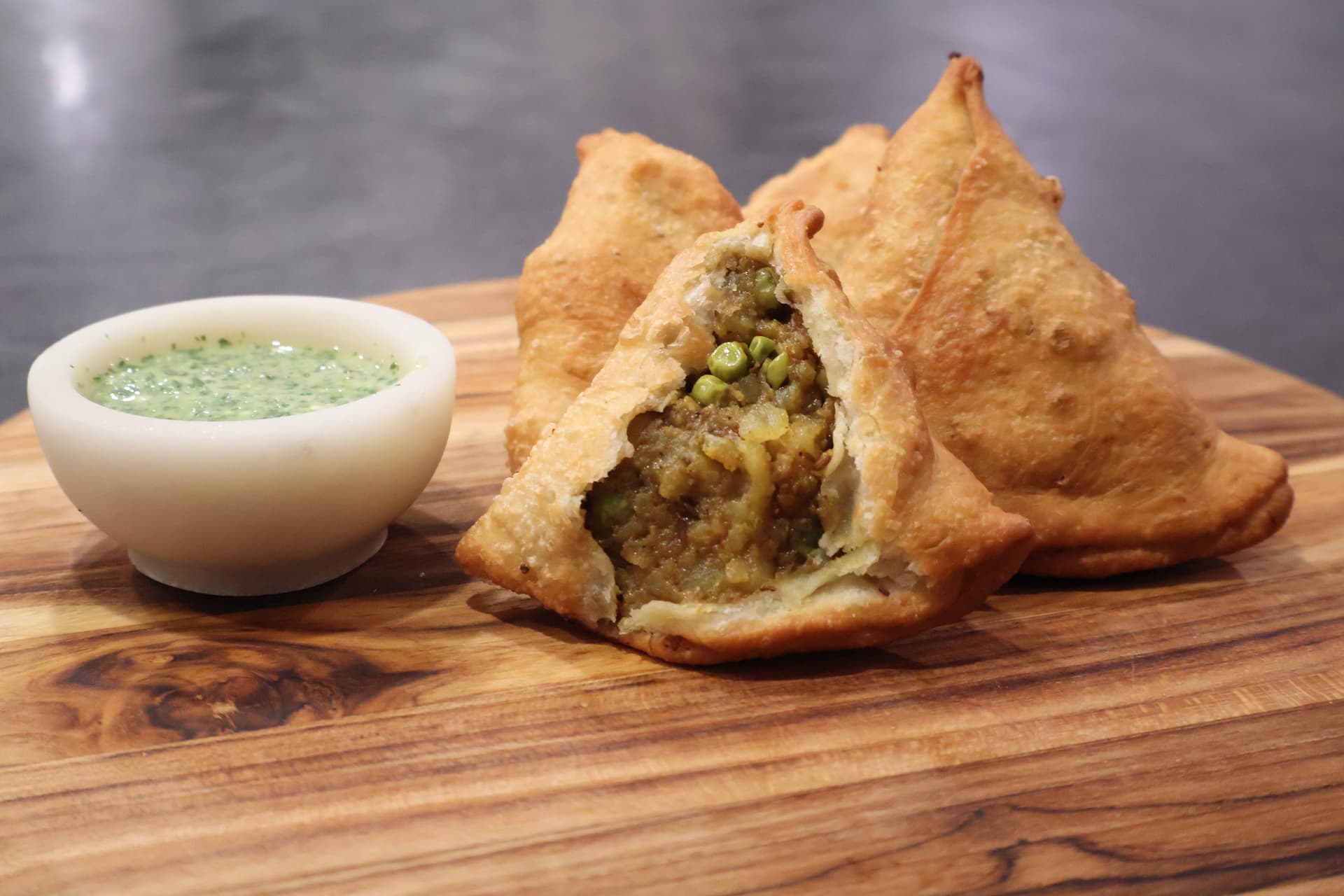 Golden crispy samosa held in hand with filling visible, surrounded by more samosas and red dipping sauce on white plate.