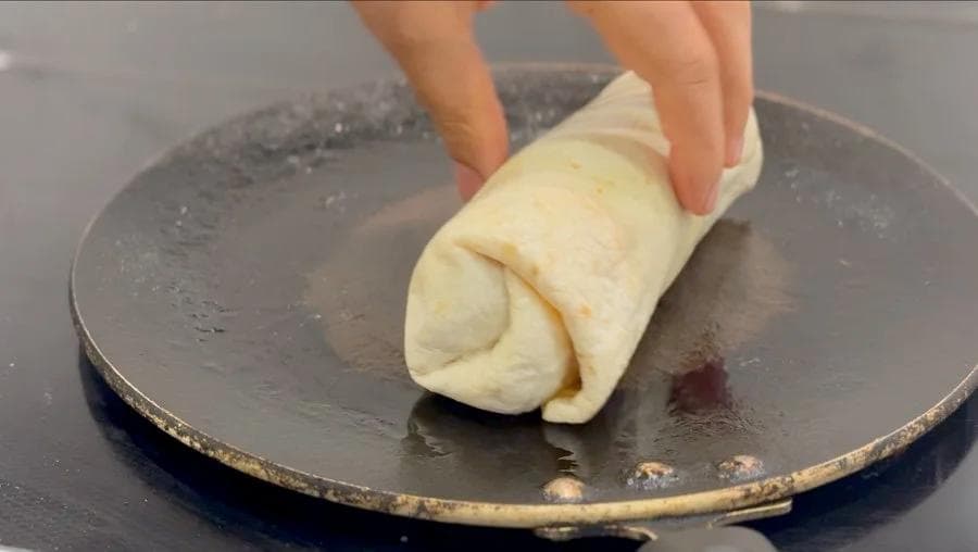 Hand placing a rolled breakfast burrito onto a dark skillet to toast.