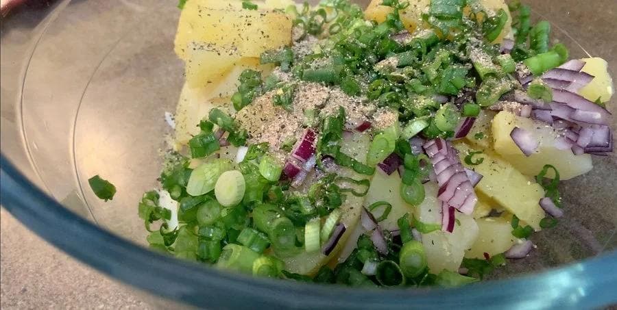 Potato salad ingredients in a bowl: diced potatoes, chopped eggs, green onions, red onions, chives, and seasonings before mixing.
