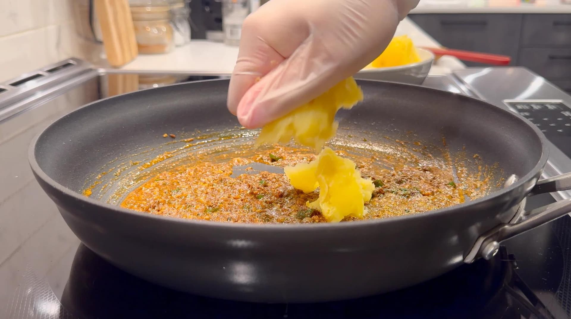 Hand adding yellow bell peppers to spiced potato mixture sizzling in a dark pan on stovetop.